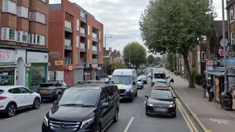 Google A Google Streetview image of Lea Bridge Road, with traffic, with shops on one side of the road and a bus stop on the other.