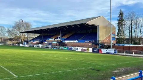 Martin Giles/BBC A main stand at King's Lynn Town is in the centre of the frame. It has rows of blue and yellow seats. There is a large football pitch in the foreground which has white lines painted on it.
