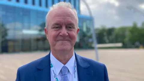 Martin Griffiths standing outside the Corby Cube building wearing a blue suit, white shirt and blue tie.
