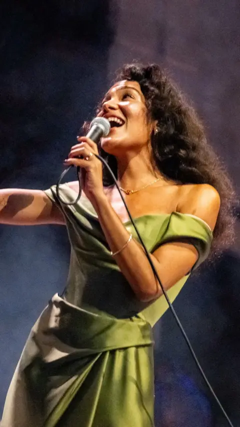 Olivia Dean wearing a green dress, holding a wired microphone while performing on stage. 