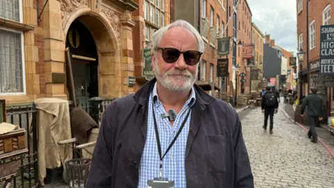 A man in sunglasses standing on a cobbled street made up to look like Victorian London with old signs and street furniture. He is wearing a blue and white shirt with a grey jacket and lanyard. He has a short beard and grey hair. Behind him, two men can be seen walking away with their backs to the camera.