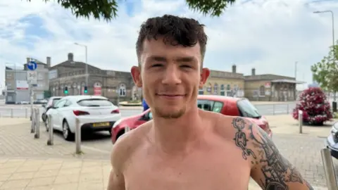 Andrew Turner/BBC Bailey Woollard has brown hair, which is cropped on the sides and back, and longer on the top and front. He has tattoos on his left arm, and is not wearing a top. He is pictured on the Station Square in Lowestoft, with Lowestoft Central railway station in the backdrop, with cars parked behind him.