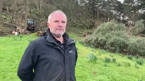 A man with a black jacket smiles at the camera , behind him you can see tree surgeons working on fallen trees 