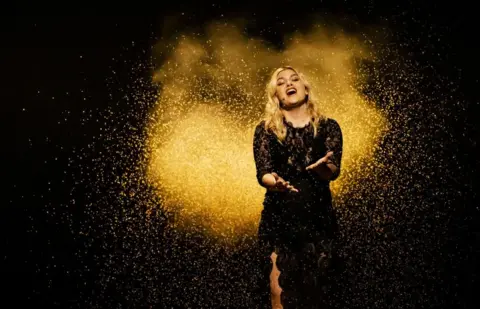 EPA-EFE/Shutterstock France's Eurovision entry Louane places her hand out as she sings with her eyes closed, wearing a black dress and surrounded by a gold confetti-like effect, with a black backdrop.