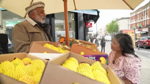 A man with a white beard and wearing a hat and beige jacket is standing behind a roadside stall in Tooting. He is holding a halved mango, while a customer looks on. Boxes of mangoes fill the foreground and a busy street runs behind them.