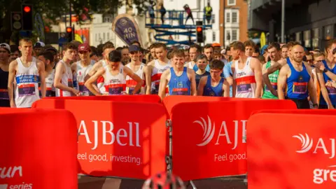 AJ Bell Great Bristol Run Male athletes standing on the road as they get ready for the start of the race. They are all wearing vests with their race bib on the front. They are standing behind some metal gates that are covered with red fabric with the AJ Bell logo.
