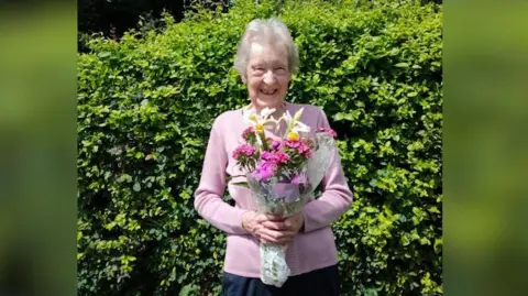 A woman stands in front of a hedge on a sunny day. She has white hair and is wearing a pink cardigan and black trousers. She is holding a bouquet of pink and white flowers and is smiling into the camera.