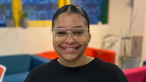Anya smiles at the camera. She wears a black jumper and glasses.