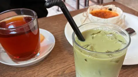 Supplied Close-up image showing a green coloured matcha latte in a glass, next to a cup of tea in a glass, resting on a white saucer, with a cheesecake dessert on a plate behind.