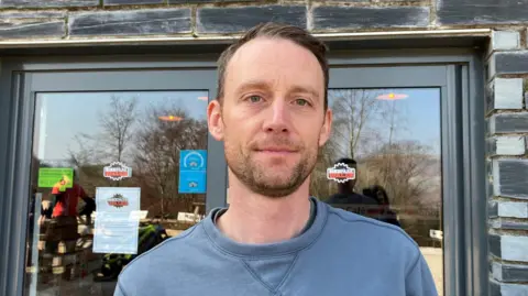 Martin Astley has brown hair and a beard wearing a blue top. He is standing outside his business premises. There is a glass door in the background with a hygiene rating on it, as well as stickers with the bike park logo on and a white piece of paper attached to it.