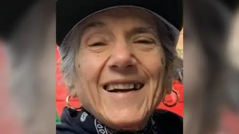 Pepita Sakirecili, also known as Pepita Garcia, wearing a cap and gold hoop earrings laughing toward the camera. She is wearing a patterned scarf and dark top, and has grey curly hair, either worn short or tied back.