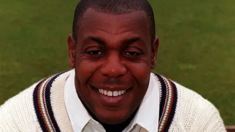 PA Media David 'Syd' Lawrence. He has short dark hair and is wearing a white collared shirt under a cream knitted jumper. He is looking directly at the camera and smiling 