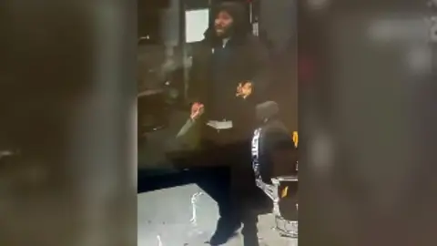 A CCTV still shows a man in a black coat with his hood up standing in a barber's shop holding a long kitchen knife in his right hand.