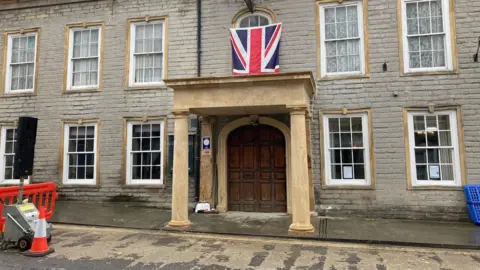 Four sand-coloured pillars are holding up a stone balcony above a big wooden door to a grey-stoned building which is filling the image. 