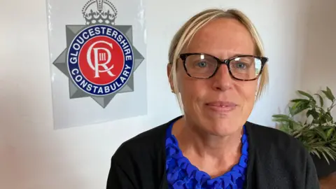 A white woman is looking at the camera with a closed smile. She is wearing brown framed glasses and has blonde hair tied back. She is wearing a black cardigan and blue top. A sign that says Gloucestershire Constabulary is on the wall behind her to the left of the photograph. A potted plant is to the right.