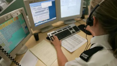 A woman with her hair pulled back into a ponytail sits at a desk with three screens and a keyboard. She has a headset on and is wearing a white blouse with a black epaulette labelled "ENQUIRY OFFICER" in white lettering.