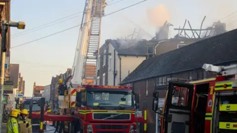 Fire engines and firefighters in a road of shops and flats above with smoke seen coming from the roof of a building.