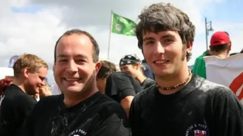 RNLI Will and his father are smiling at the camera. Will looks like he is in his late teens and they are both wearing black RNLI-branded T-shirts. His father has short brown hair, while Will has sideburns and is wearing a necklace. There are people behind them a flag flying. 