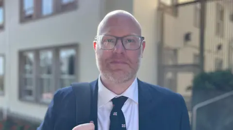 Deputy Rob Curgenven - A man with a bald head, wearing black rimmed glasses, with a ginger and grey beard. He is wearing a blue suit and a white colleared shirt, with a navy blue tie, with Guernsey flags on it. He his holding a navybag strap over his right shoulder. 