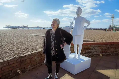 Penguin Random House Wilson standing by a sunny Brighton beach in front of a white statue of herself covered in tattoos, leaning on books. 