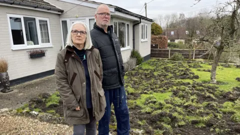 Edd Smith/BBC Lorraine and Phil Williams stood by their damaged front lawn. Lorraine, who has shoulder-length white hair and black glasses, is wearing a brown wax jacket. While Phil, who has short grey hair and thin glasses, is wearing a black puffer jacket and jeans.