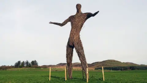 Rab McMurdo A large wicker sculpture of a man with his arms held out, standing in the middle of a grass field