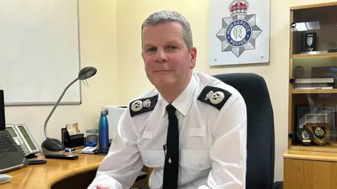 BBC Humberside Police Chief Constable Chris Todd. He has short brown hair and is sitting in his office in a white uniform shirt with black epaulettes with a crown and badge on and a black tie with the force's badge tie pin