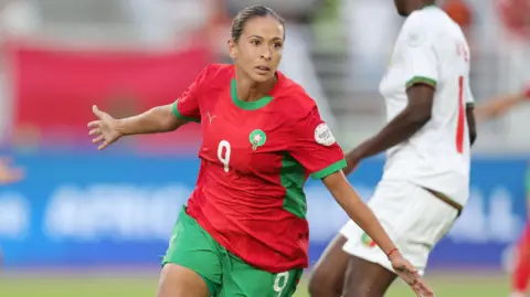 Ibtissam Jraidi in red Morocco shirt and green shorts spreads her arms to her sides and wheels away in celebration as a Mali player in white is seen in the background