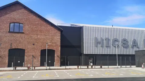 The Housing Innovation and Construction Skills Academy (HICSA) at Riverside Sunderland. There is a modern-looking brick building next to an office building which has modern glass panels on it. HICSA reads in large letters. 