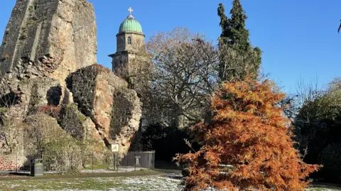 A church building and large brick ruin are in the background, surrounded by trees, along with an iron fence. In the foreground is grass covered in snow, as well as a tree with orange leaves.