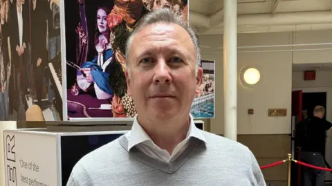 LIPA principal and chief executive Professor Sean McNamara has grey hair and wears a white shirt with a grey v-neck jumper. He stands in the LIPA building in front of a sign.