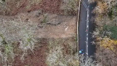 Westmorland and Furness Council An aerial view of the muddy landslip area - to the right the narrow road is visible with extensive crash barriers in place. 