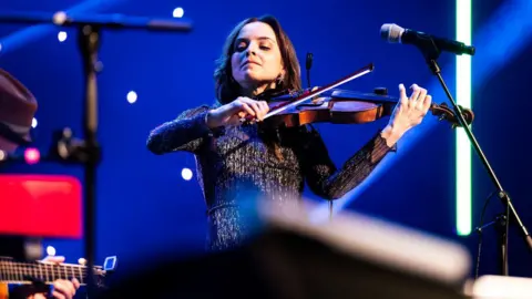 Paul Campbell A fiddler plays her instrument as she stands next to a microphone and against a blue backdrop.