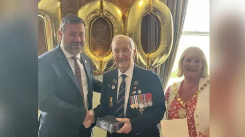 Image depicting councillor Kevin Guy in a navy suit and red tie, shaking hands with D-Day Veteran Stan Ford who is wearing his military regalia and holding a gift received from the council. Also pictured is councillor Karen Walker who is wearing a white waistcoat, red and white top and white trousers.