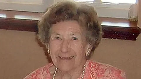 A woman with light-brown hair smiling. She is wearing a pink top, a pearl necklace and matching earrings. Behind her is a wooden windowsill.