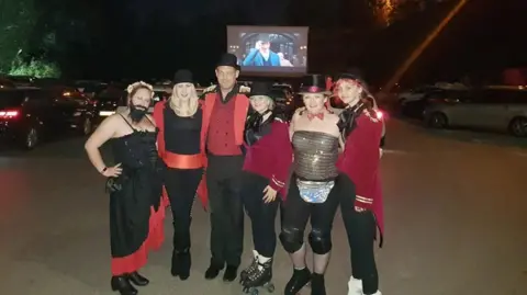 Karen Hall/Facebook A group of people stand at the drive in cinema, dressed in The Greatest Showman attire. 