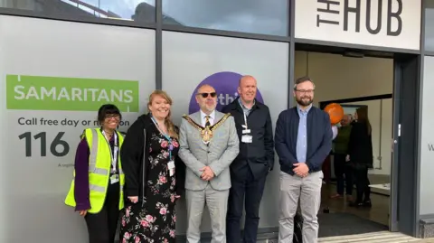 Five people, including Kirsten Rose and the Wolverhampton mayor, stand outside the glass frontage of The Hub at the station. One woman works for WMR and has a florescent bib over her clothing. The mayor is wearing a suit with his mayoral chains on top with two other men in navy and grey casual clothing. Kirsten is wearing a floral dress and dark jacket.
