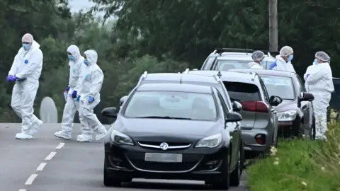 Alan Lewis A number of people dress in white crime scene suits are pictured standing on a road beside a uniformed police officer. They all have their faces covered with blue masks. Beside them is a line of grey cars.