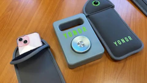 BBC Two grey neoprene phone case sits on a brown wooden table. A phone with a pink case can be seen inside one. A metal magnet remover sits in between.  It has the word 'YONDR' printed on the front in green text.