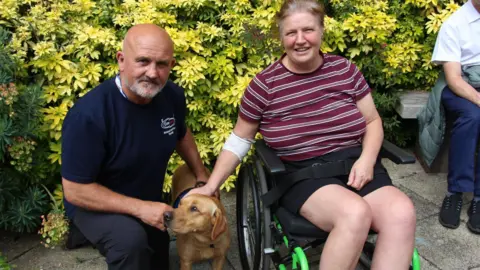 South Tees Hospitals NHS Foundation Trust A bald man crouched on the left of Kyro. He is looking into the camera with a slight smile and has a grey beard. He is wearing a navy blue t-shirt and trousers. On the right, Kyro is looking to the left. Elizabeth Donlan is on the right in a wheelchair and is smiling into the camera with grey and brown hair which is tied back. She is wearing a maroon striped t-shirt and black shorts. There is a bush behind them with yellow and green leaves.