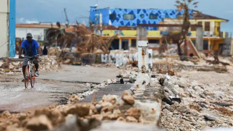 A man rides a bicycle along a concrete road, past damaged buildings in the aftermath of Hurricane Melissa, in Black River, Jamaica,