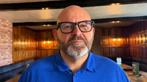 A man with a bald head and salt and pepper beard. He is wearing a blue shirt and black glasses. He is standing in his pub, which has wooden tables and a mixture of wood-panelled walls and exposed brickwork. Lighting on the side of the walls casts a golden hue.