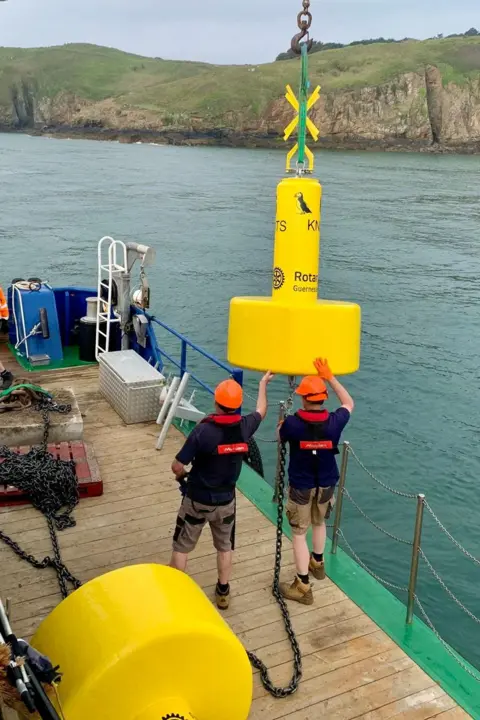 A large yellow marker buoy is being winched into the air from a boat and two men wearing hard hats are helping to guide it. There is another marker buoy lying on the deck. The shore of Herm is visible across the sea.