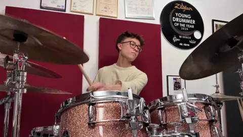 Ellis Gates wearing glasses and a white jumper playing the drums