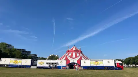 Set against a blue sky is a red and white striped big top with three trailers branded with Gandey's parked in front.