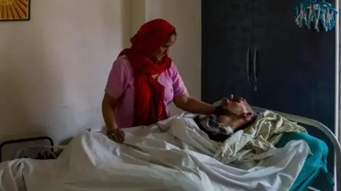 BBC/Rana Family Harish Rana's mother, wearing a pink tunic and a scarf over her head, pats her son who is lying in a bed, covered by blankets.