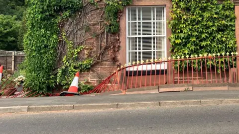 Roadside shot. It shows a stone building covered with green ivy. A large white-framed window sits above a red-painted metal railing which is bent and collapsing along the pavement. An orange-and-white traffic cone lies beside the damaged railing. 