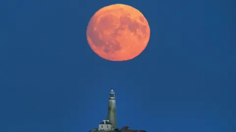 A red-orange full Moon hangs low in the night sky over a lighthouse