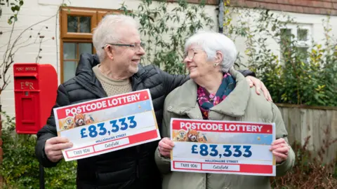 Lotería Popular de Códigos Postales. Un hombre mayor abraza a una mujer mayor. La pareja se sonríe sosteniendo billetes de lotería con £83,333 cada uno.