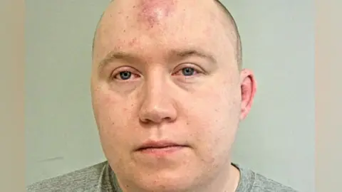 Jacob Hill's mugshot. He is a man with receding, cropped light brown hair. He is photographed in a grey t-shirt.